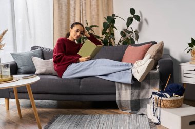 Genç bir Afrikalı Amerikalı kadın koltuğunda oturup şık, modern dairesinde kitap okumaktan zevk alıyor..