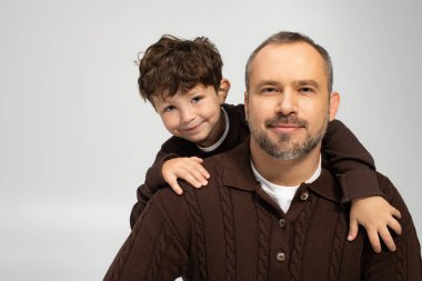 Neşeli küçük bir çocuk babasının omuzlarına sarılıyor. Sıcak bir ortamda sıcak bir gülümseme paylaşıyor..