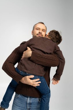 Bir baba, genç oğlunu sevgiyle kucağına alır. Her ikisi de rahat kıyafetleriyle sıcacık ışık saçar..