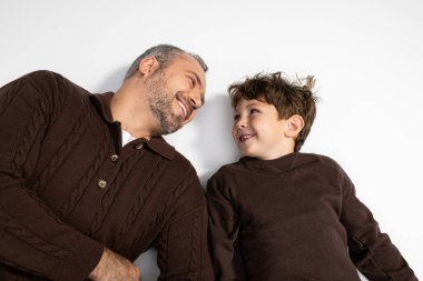 Bir adam ve küçük oğlu, yumuşak bir yüzeyde yan yana yatarken neşeli bir gülümseme paylaşıyorlar..