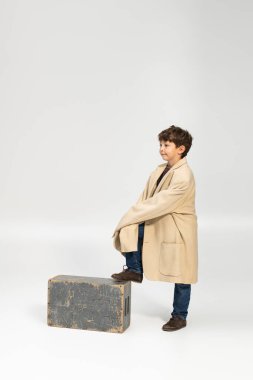 Genç bir çocuk, yüzünde neşeli bir gülümsemeyle, büyük bir palto giymiş, bir kutunun üzerinde duruyor..