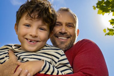 Baba ve oğul neşeli bir şekilde kucaklaşır, ikisi de güneşli bir günde gülümserler..