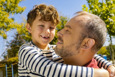 Bir baba, güneşli bir park ortamında gülümseyen neşeli küçük oğlunu kucaklar..