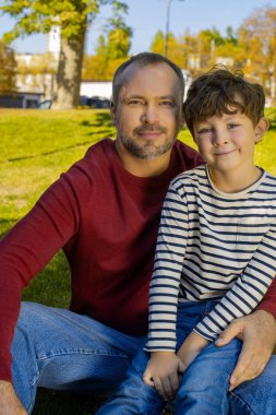 Bir baba ve küçük oğlu çimenlerin üzerinde oturup, mutlulukla dolu güneşli bir günün tadını çıkarıyorlar..