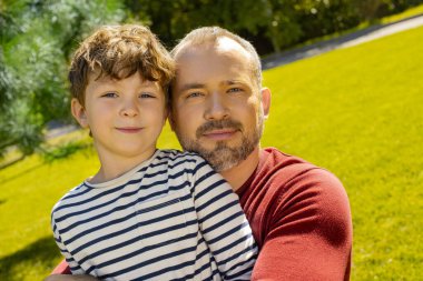 Babası küçük oğlunu canlı yeşil bir parkta tutuyor. Mutluluk ve sıcaklıkla gülümsüyor..