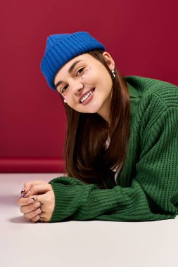 Mavi bereli neşeli bir genç kız kırmızı bir stüdyoda şık bir şekilde poz veriyor, kış görüntüsünü sergiliyor..