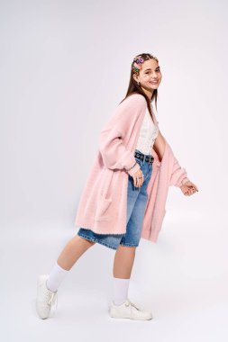 Yanakları ışıltılı şık bir kız, gri bir stüdyoda rahat pembe bir hırkayla poz veriyor..