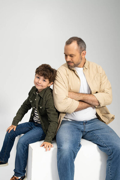 Отец сидит на белом блоке, излучая спокойствие, в то время как его сын игриво наклоняется рядом с ним, улыбаясь.