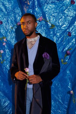 Şık bir genç adam mavi bir arka planda canlı çiçekler arasında güvenle duruyor..