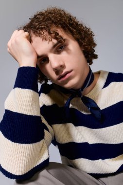 Kıvırcık saçlı bir genç şık çizgili süveter ve rahat bir duruş takınır..