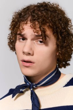 Kıvırcık saçlı ve eşarplı bir genç, basit bir arkaplanda dinamik bir şekilde poz veriyor..