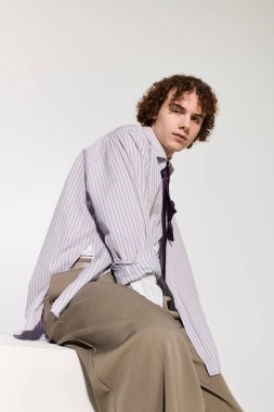 Genç, modaya uygun kıyafetlerle ve sade bir arka planda kendine güvenen bir pozla dikkat çekiyor.