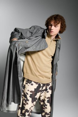 Şık giyinen genç, basit bir gri zemine karşı özgüvenle poz veriyor, enerji saçıyor..