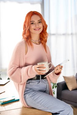 Kızıl saçlı genç bir kadın şık dairesinde, elinde sıcak bir içecek tutarak rahatlıyor..