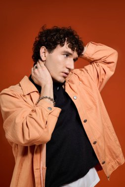 Yakışıklı bir adam kendine güvenerek poz veriyor, çarpıcı turuncu bir arka planda şık kıyafetinin altını çiziyor..