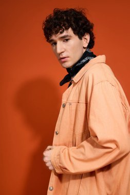 Handsome man in trendy attire stands poised against a bright orange wall, exuding charm and style.