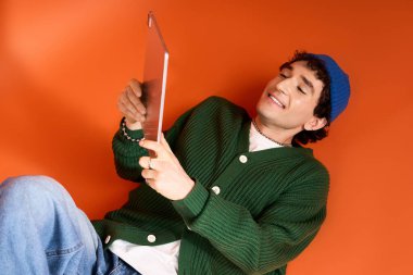 Man in green outfit and blue beanie smiles at his tablet in a vibrant setting.