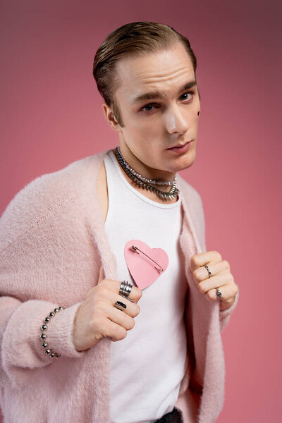 A confident man shows off a playful pink heart pin against a soft pink backdrop.