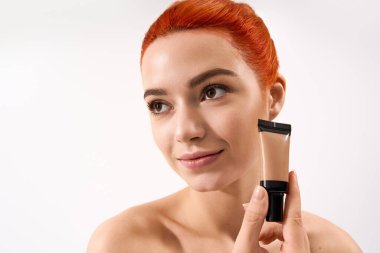 Kızıl saçlı genç bir kadın makyaj malzemesi tutuyor, doğal ışıkta hafifçe gülümsüyor..