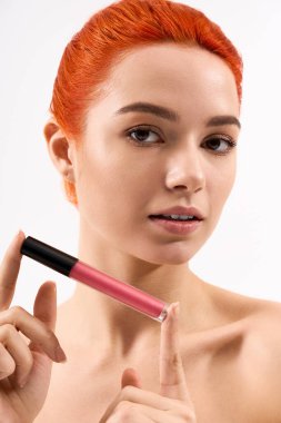Çarpıcı kızıl saçlı güzel bir kadın bir dudak ürünü tutuyor, şık makyaj seçimini sergiliyor..