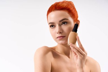 Canlı kızıl saçlı genç bir kadın makyajını parlatıyor, temeli parlak bir ışıkla tutuyor..