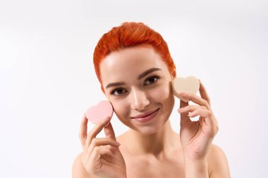 Neşeli genç kadın pembe ve krem rengi kalp şeklindeki makyaj malzemelerini tutuyor, özgüvenle gülümsüyor..