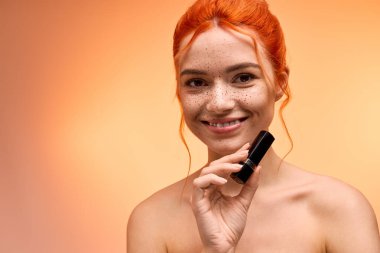 Kızıl saçlı genç bir kadın gülümsüyor, elinde turuncu bir arka planda güzellik ürünü tutuyor..