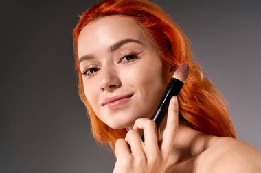 Turuncu saçlı güzel bir kadın makyaj yeteneklerini sergilerken yumuşak ışıkta zarif bir şekilde gülümsüyor..