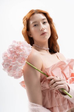 Canlı kızıl saçlı genç bir kadın elinde pembe çiçeklerle stüdyoda zarifçe poz veriyor..