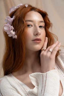 Uçan kızıl saçlı genç bir kadın stüdyoda bahar çiçeklerinin yanında güzellik ve zarafet yayar..