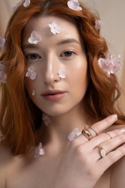 Kızıl saçlı genç bir kadın minimalist stüdyo ortamında çiçeklerle zarif poz veriyor..