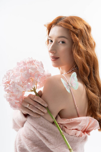 Redhead woman poses gracefully with pink flowers, showcasing trendy spring fashion in a studio.