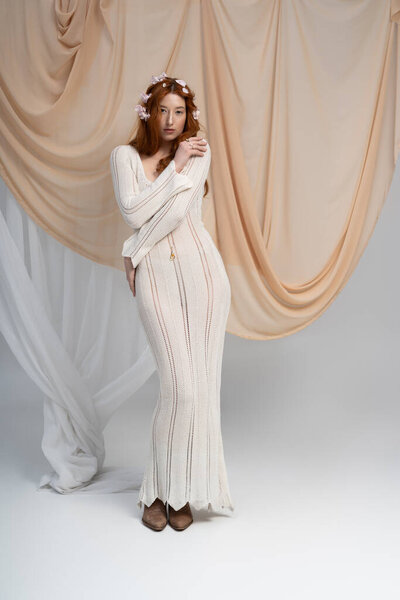 A young redhead showcases a stylish outfit among delicate flowers in a calm studio.