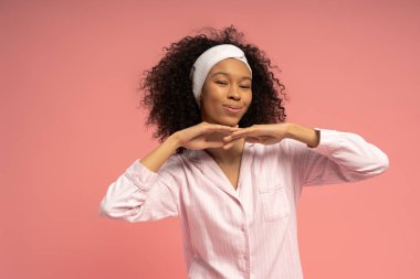 Kıvırcık saçlı genç bir kadın pembe bir ortamda şakacı bir poz verirken ışıl ışıl gülümser..