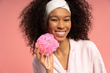 Kıvırcık saçlı neşeli kadın pembe saç tokasını yumuşak pembe bir fonda tutuyor..