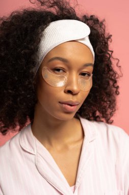 Kıvırcık saçlı genç bir kadın şık bir göz bandı takarken gençleştirici bir güzellik rutininden hoşlanıyor..