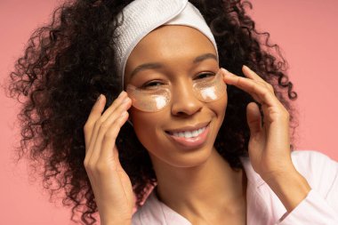 Genç bir kadın göz bandı altında başvuruyor, evde neşeli bir gülümsemeyle kendine bakmaktan zevk alıyor..