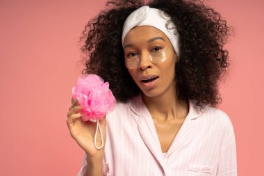 Kıvırcık saçlı genç bir kadın pembe banyo aksesuarlarıyla cilt bakımını yapıyor..