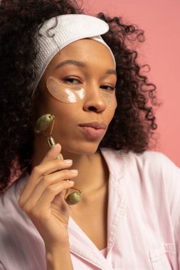 Kıvırcık saçlı güzel genç bir kadın cilt bakımının bir parçası olarak yeşim bir silindir kullanıyor..