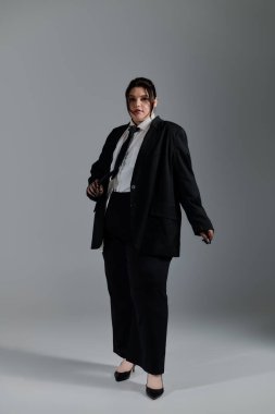 Gösterişli siyah takım elbiseli kendine güvenen, iri yarı bir kadın basit bir arkaplana karşı şık bir şekilde poz verir..