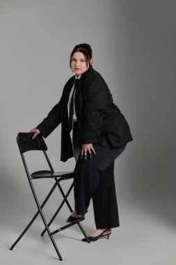 Çekici ve cüsseli bir kadın, özel dikim bir takım elbiseyle koltuğun yanında rahatça poz veriyor..