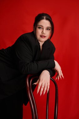 Artı boyut, zarif bir poz verir. Kalın kırmızı bir sandalyeyle poz verirken..