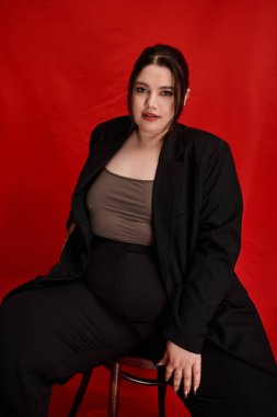 Çarpıcı, iri yarı bir kadın, tarzını koyu siyah bir elbiseyle kırmızı bir arkaplanda sergiliyor..