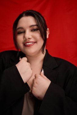 Büyük beden bir kadın şık bir elbiseyle parlak kırmızı bir arka fona sıcak bir şekilde gülümser..