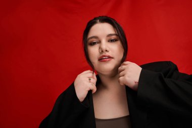 Güzel ve iri bir kadın şık poz veriyor, ceketini koyu kırmızı arka planda ayarlıyor..