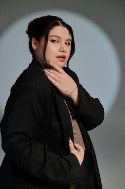 Kendine güveni tam, kilolu bir kadın şık bir elbiseyle arkasında yumuşak bir spot ışığı ile zarif bir şekilde duruyor..