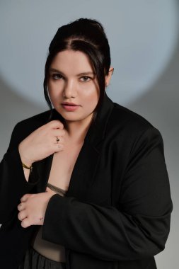 Güzel, iri yarı bir kadın zarif poz veriyor, şık siyah bir ceket giyiyor ve kendine güvenen bir ifade takınıyor..