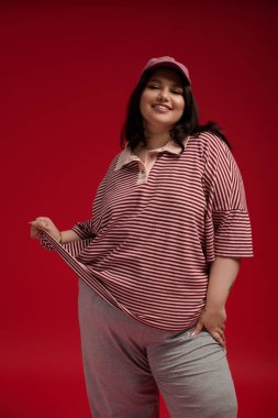 Artı boyut modeli, modaya uygun çizgili bir üst ve spor şapkaya güveni yansıtıyor, neşeyle poz veriyor..
