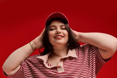 Artı kendine güveni olan cüsseli bir kadın çarpıcı bir kırmızı arka plana karşı şakacı bir şekilde poz veriyor..