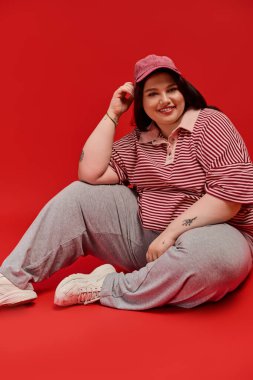 Kendine güvenen, kilolu bir kadın, şık görünüşünü ve kırmızı arka planda neşeli gülüşünü sergiler..